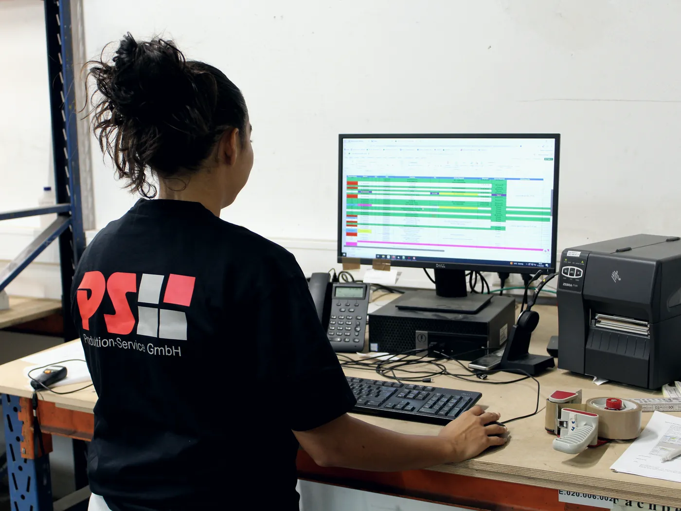 Mitarbeiterin arbeitet am Bildschirm mit Auftragsübersicht, daneben ein Etikettendrucker