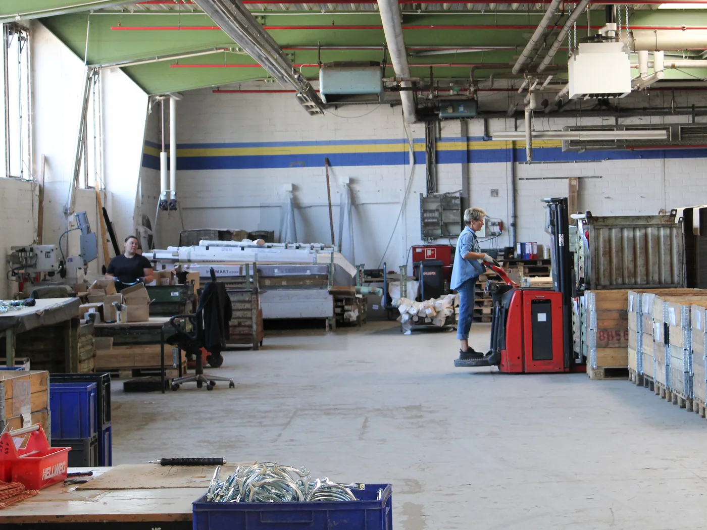 Blick in die Halle des Industrieservice: Arbeitsplätze mit Kleinteilen und Gitterboxen, im Hintergrund eine Mitarbeiterin mit Elektro-Hubwagen