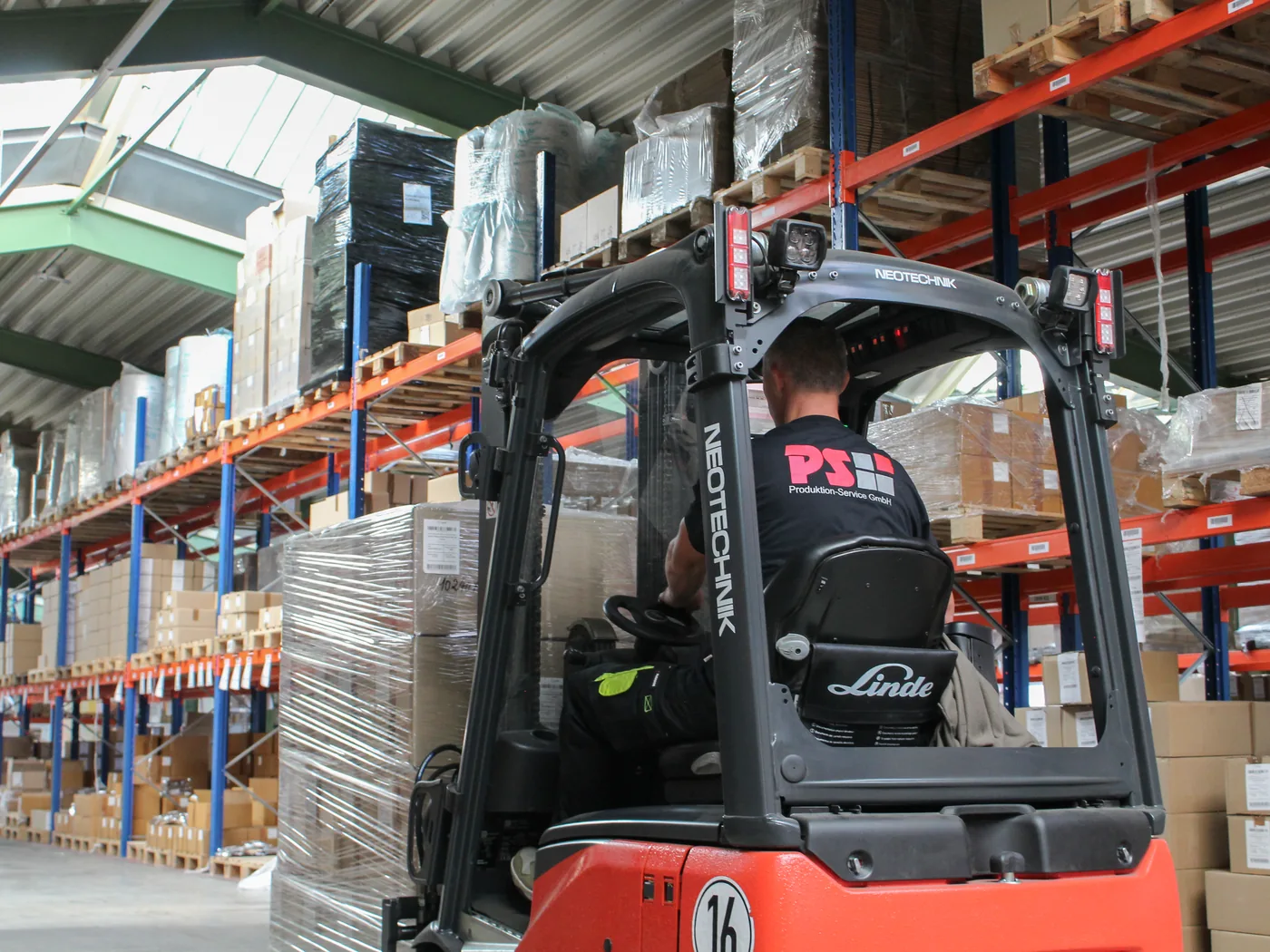 Employee in a PS shirt moving a wrapped pallet through the racking aisle on a forklift