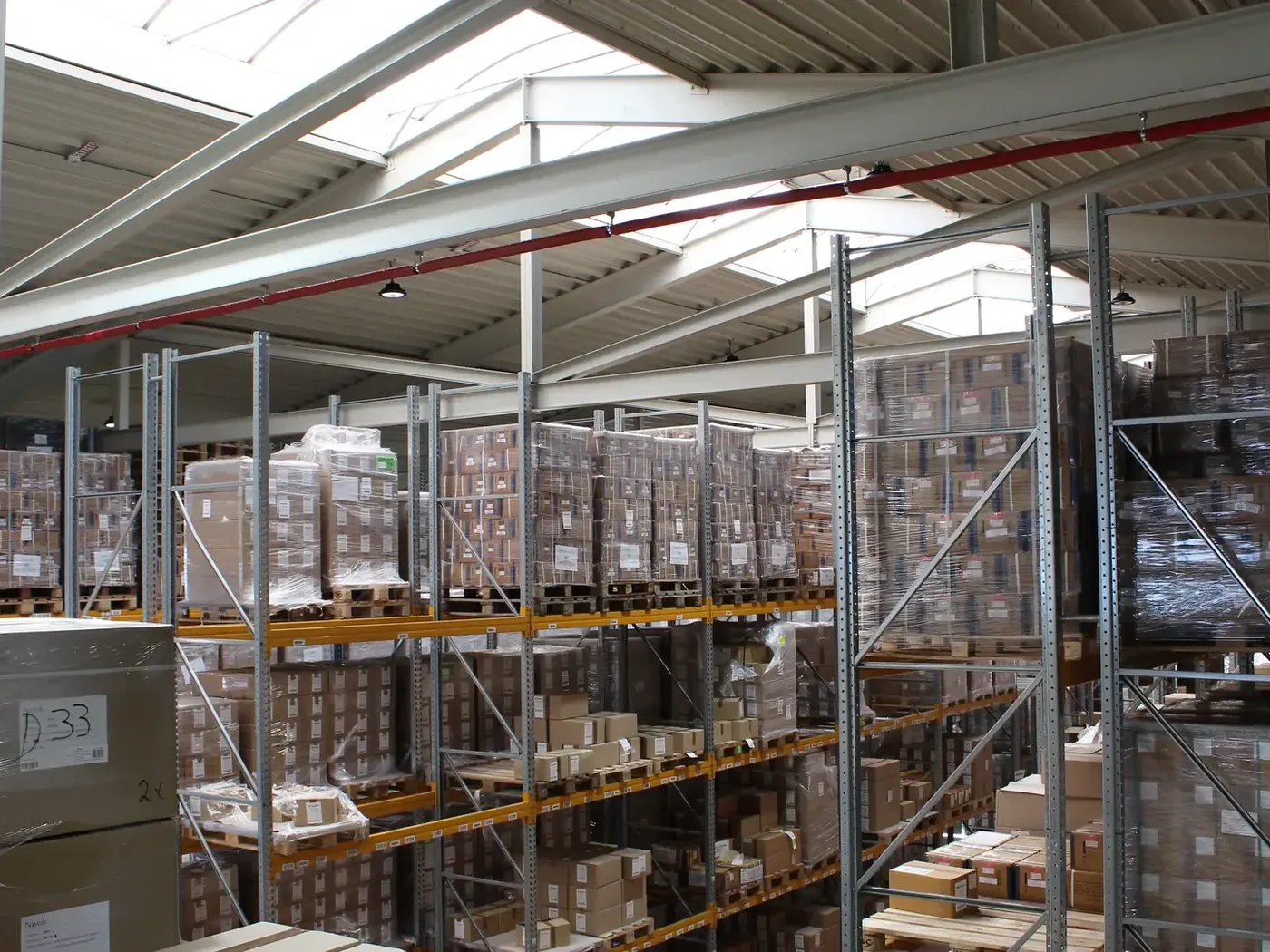 Blick durch eine Lagerhalle der PS Produktion-Service GmbH mit mehrgeschossigen Palettenregalen, gestapelten Kartons und Tageslicht aus dem Hallendach